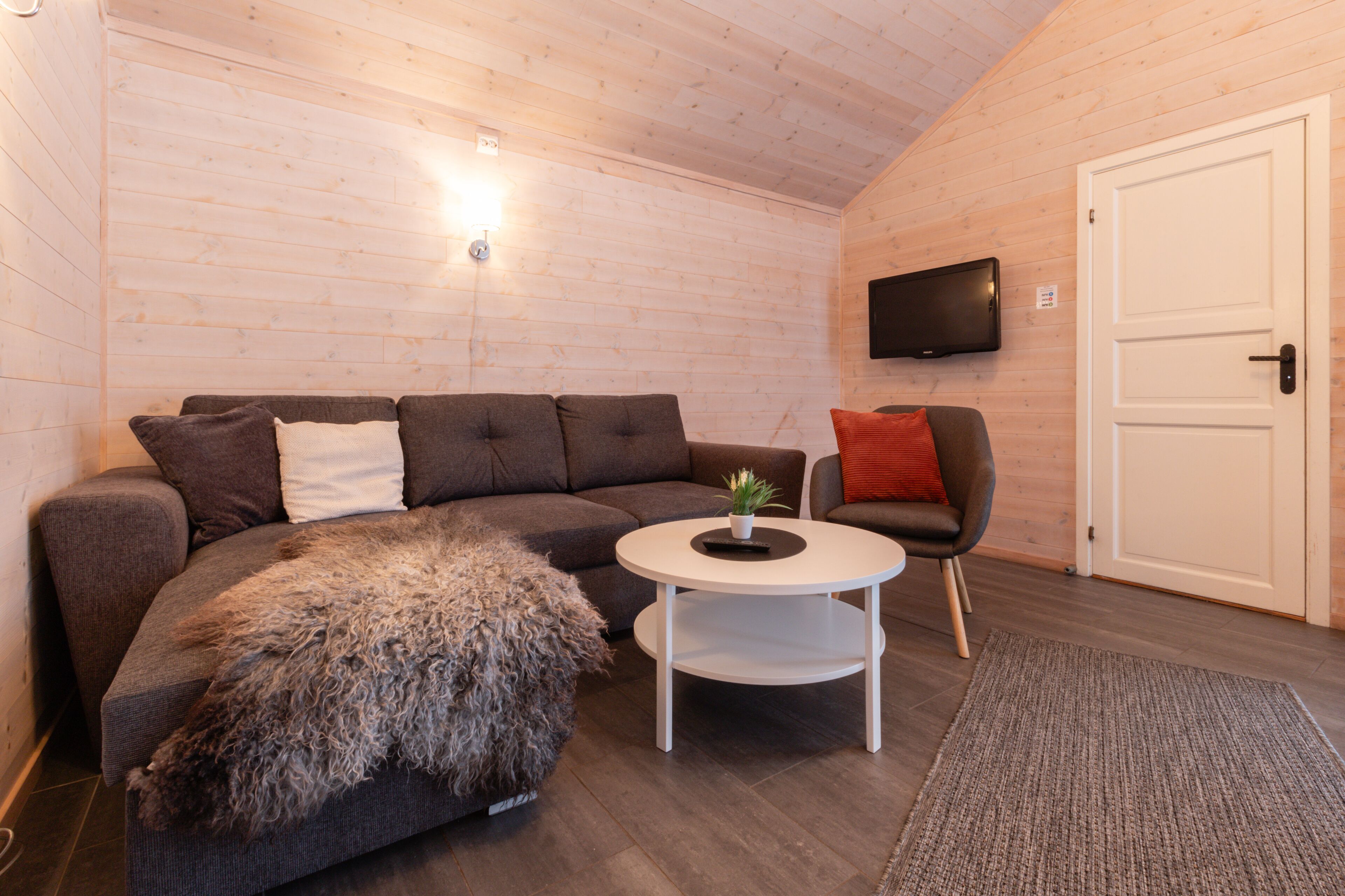 Une pièce chaleureuse avec un canapé gris aux lignes arrondies, une table basse blanche, un pouf moelleux, un fauteuil rouge et un téléviseur fixé au mur.