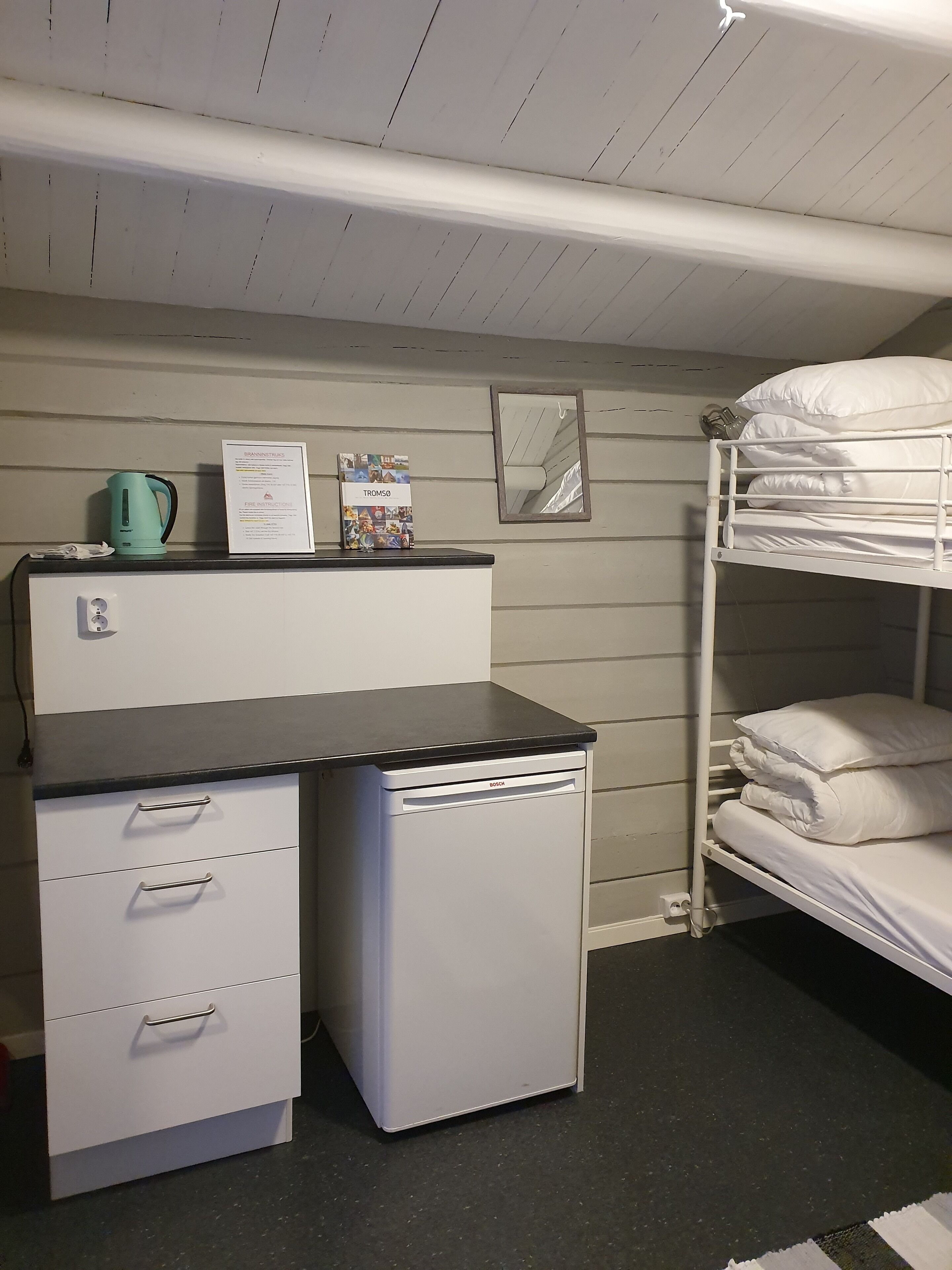 A bunk bed with white bedding, a small kitchenette with a Bosch refrigerator, a kettle, and a fire safety instruction sign.
