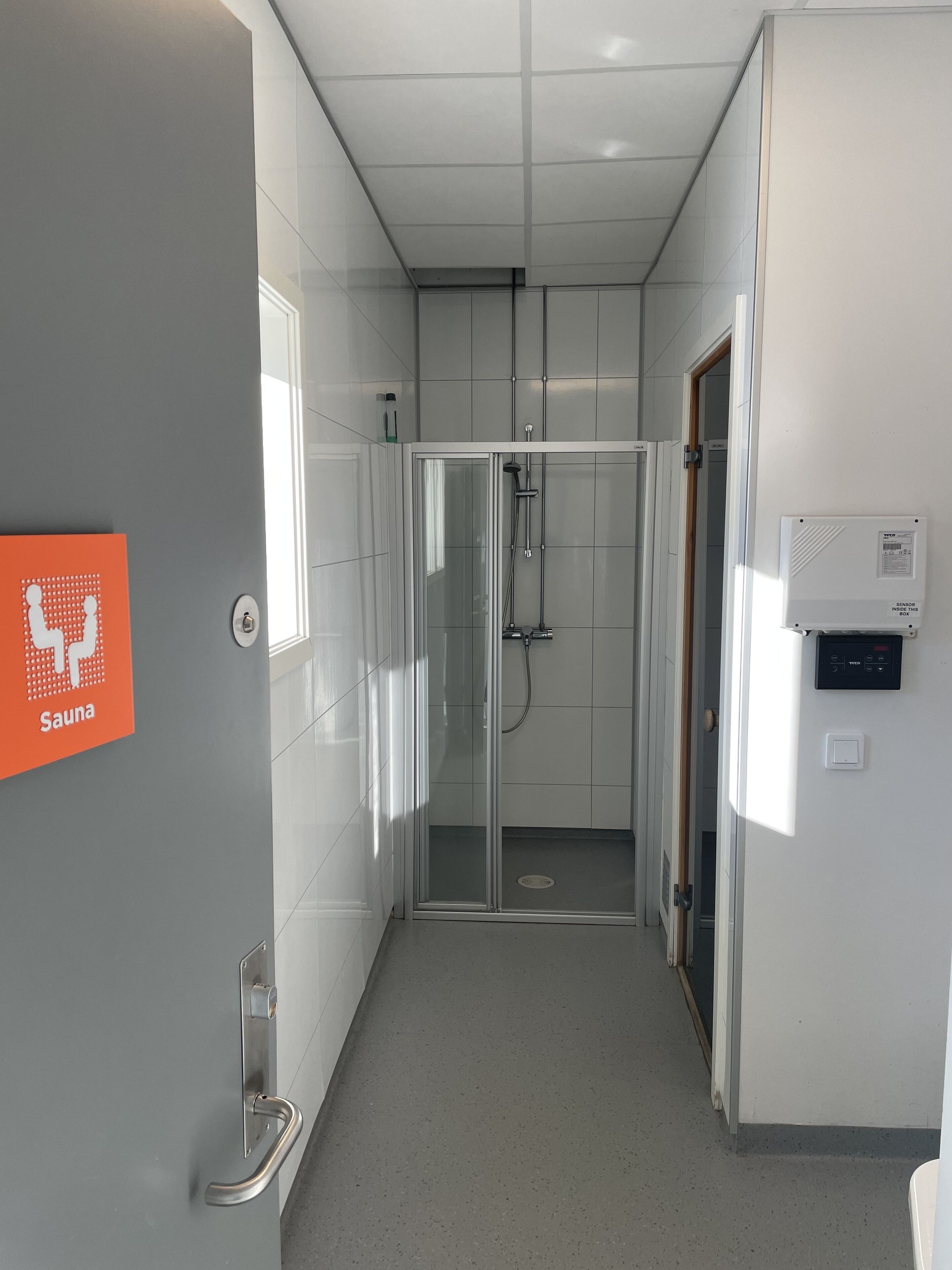 Un bany modern amb dutxa de vidre, senyal de sauna i una caixa de sensors a la paret.
