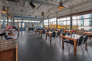 A spacious dining area with multiple tables and chairs, large windows offering views of the city, and a high ceiling with exposed beams. - Stayokay Eindhoven - Hostel (Eindhoven)