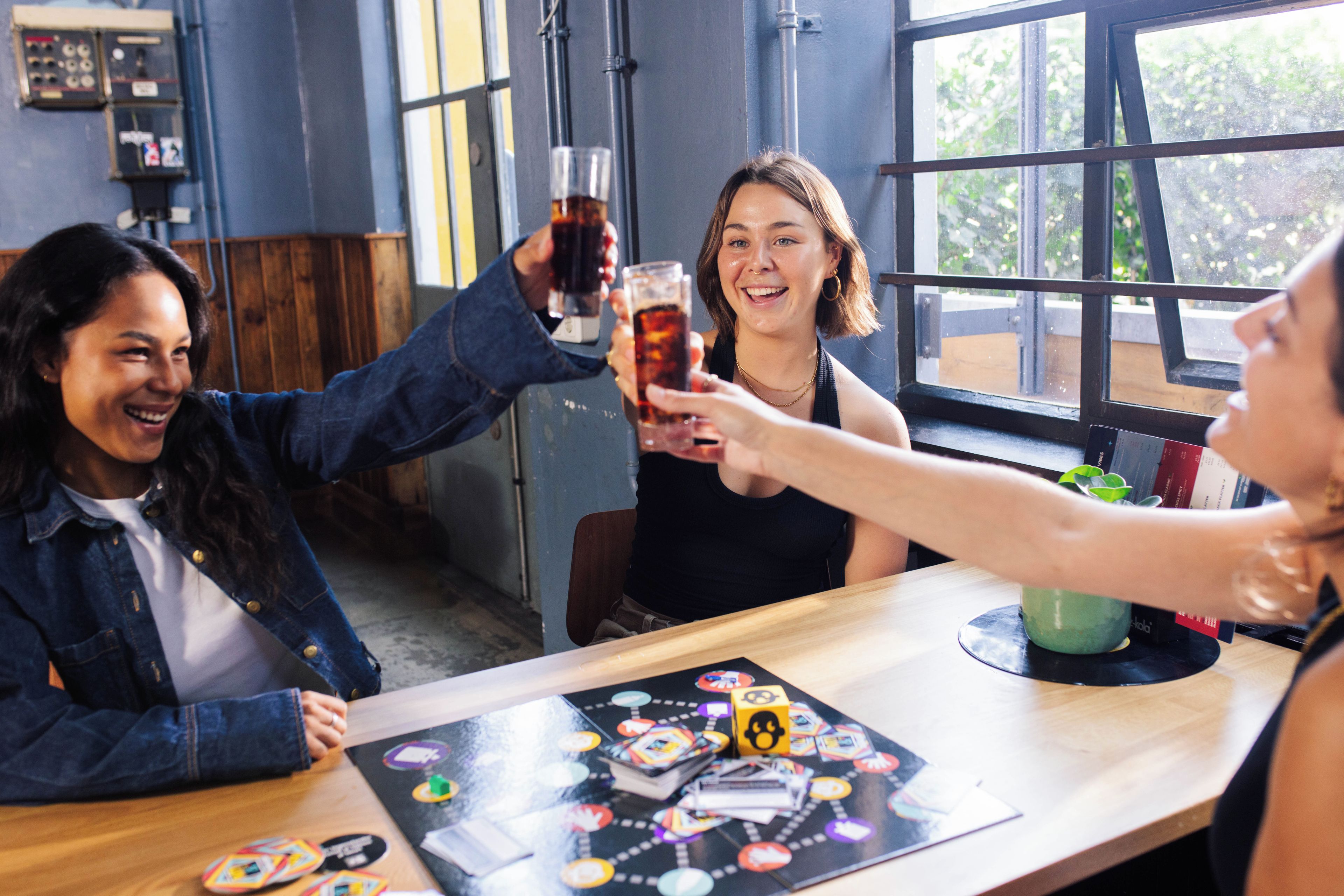 Un groupe de personnes trinquant avec des boissons autour d’une table où est installé un jeu de société.