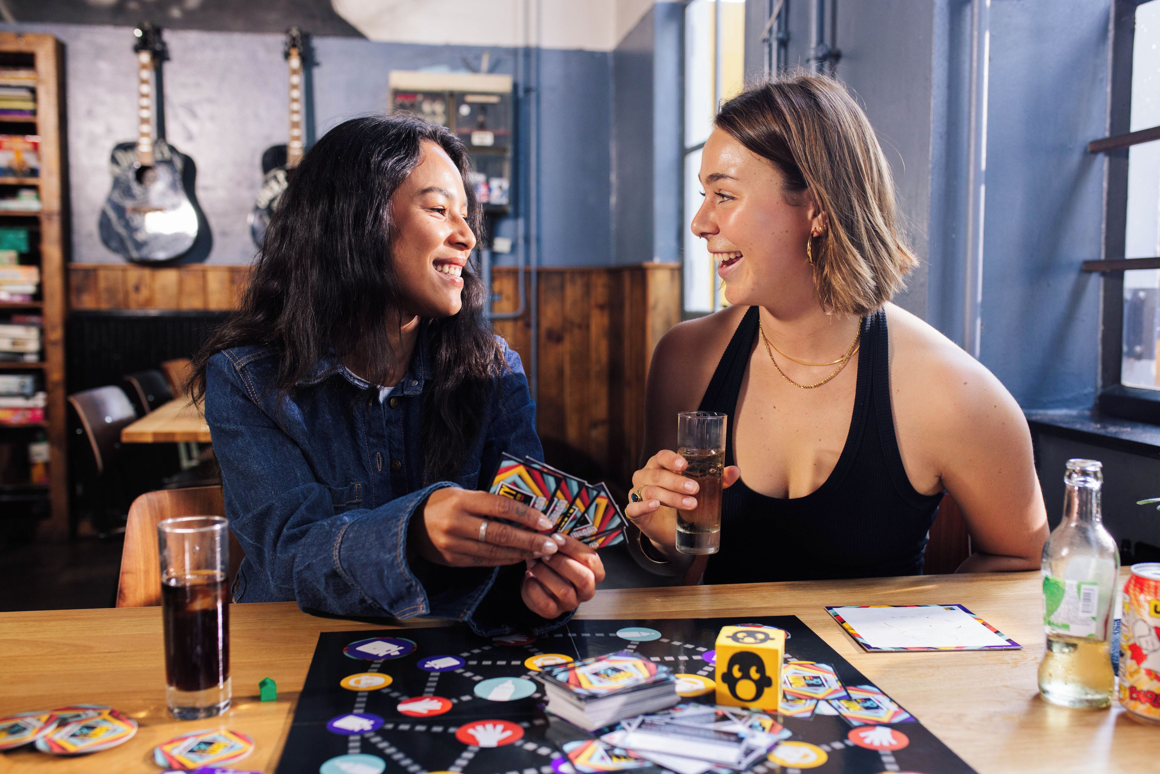 Two individuals playing a board game at a table with a drink and snacks.