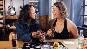 Two individuals playing a board game at a table with a drink and snacks.