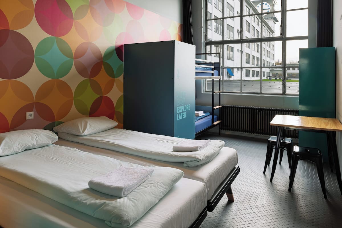 A hotel room with two single beds, a large window with a view of buildings, a radiator, and a desk with a chair.
