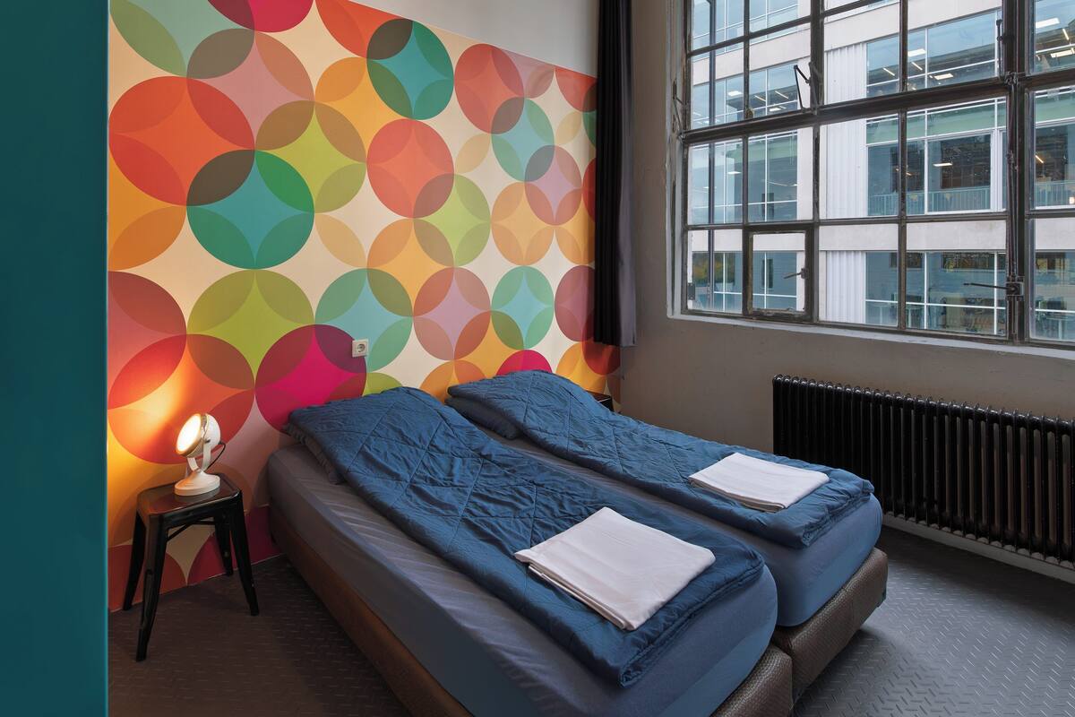A hotel room with two beds, a colorful wall, a window, and a radiator.
