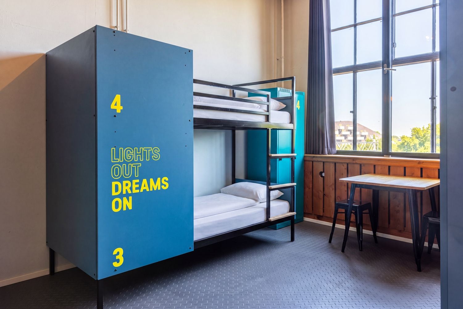 Una cama litera con un armario azul etiquetado con los números '4' y '3', y la inscripción 'LUCES APAGADAS, SUEÑOS ENCENDIDOS'.