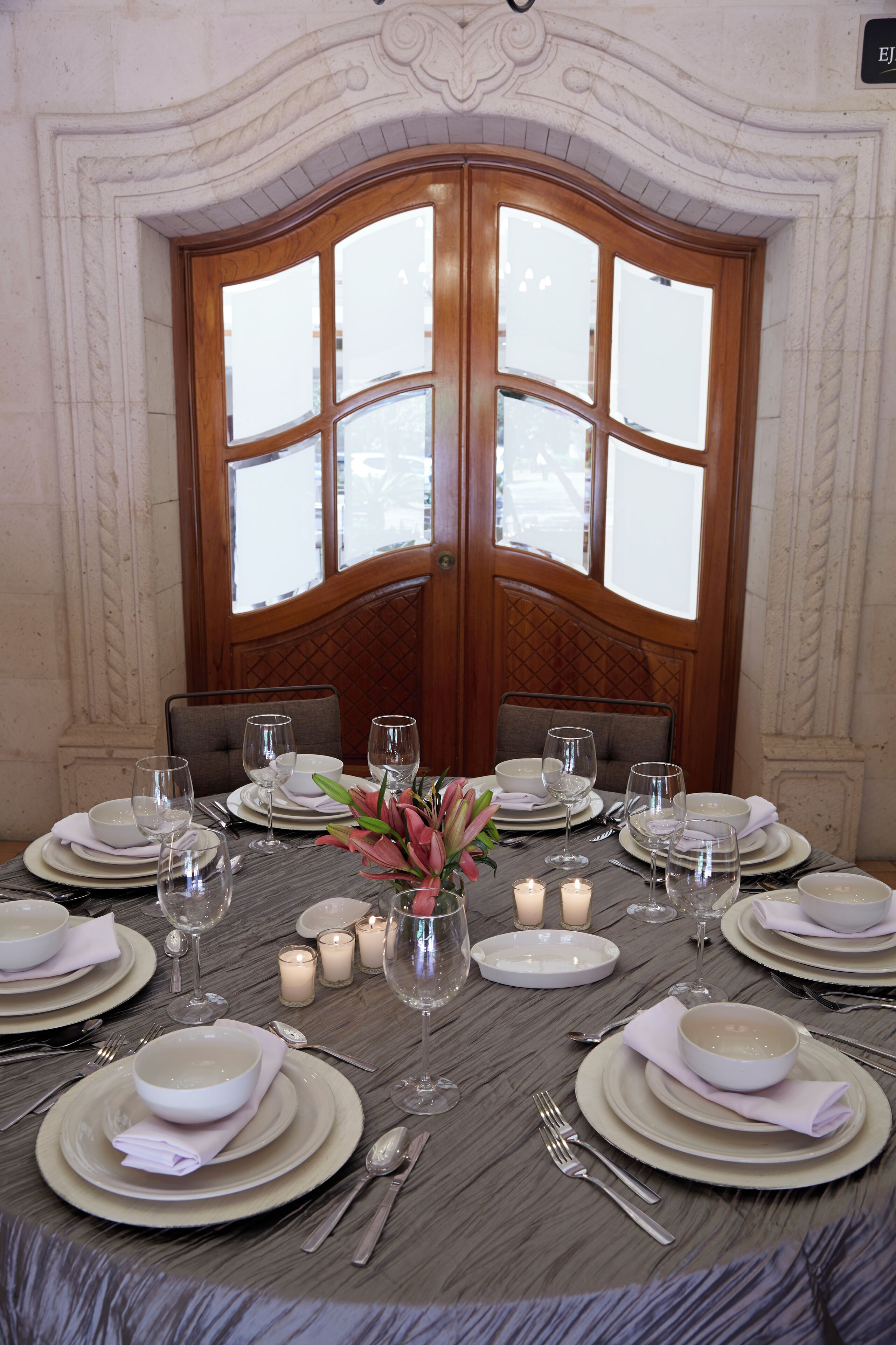 Una mesa de comedor preparada para una comida formal, con platos, fuentes y cubiertos blancos, rodeada de velas y un centro de mesa floral.