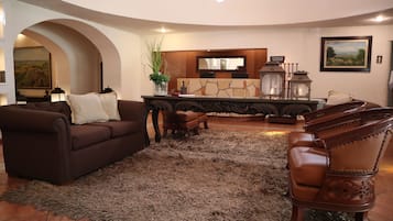 Amplia sala de estar con un sofá marrón, una silla de madera y una alfombra. Se aprecia una pared de piedra con un espejo y un cartel encima.