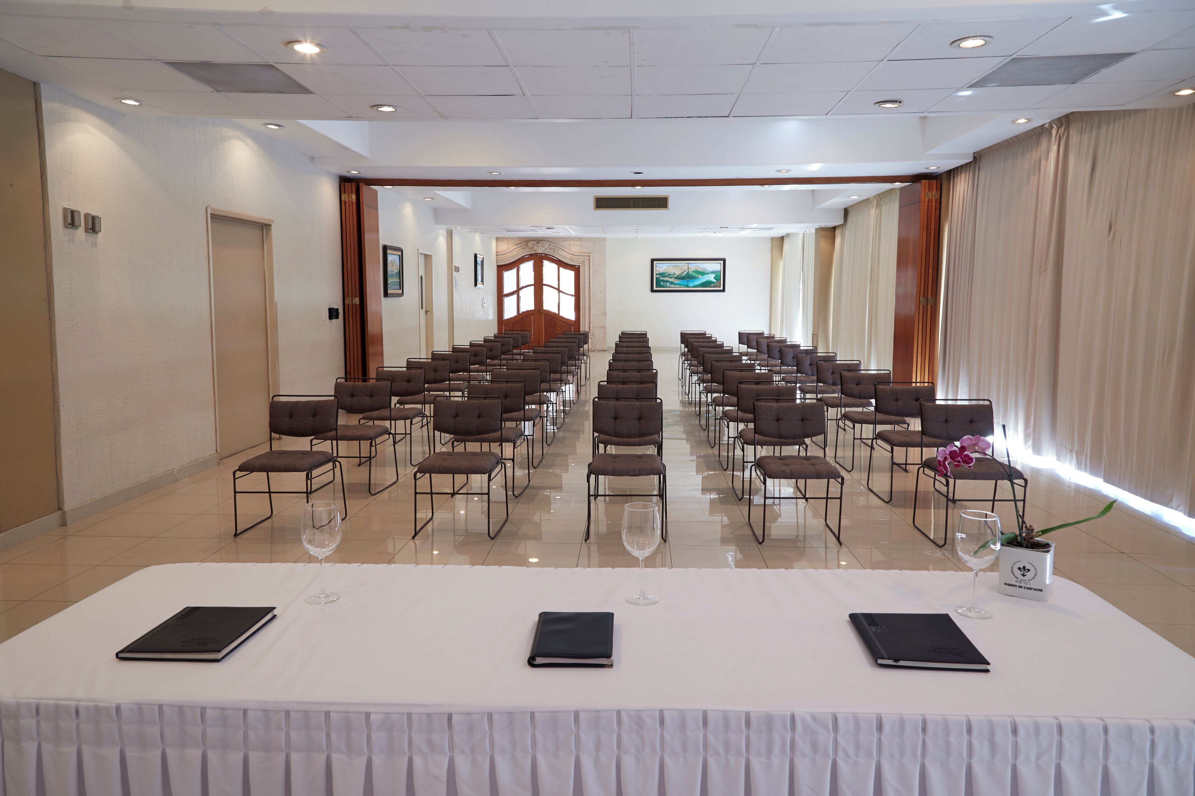A conference room with rows of chairs, a long table with a white tablecloth, and a large window with curtains.