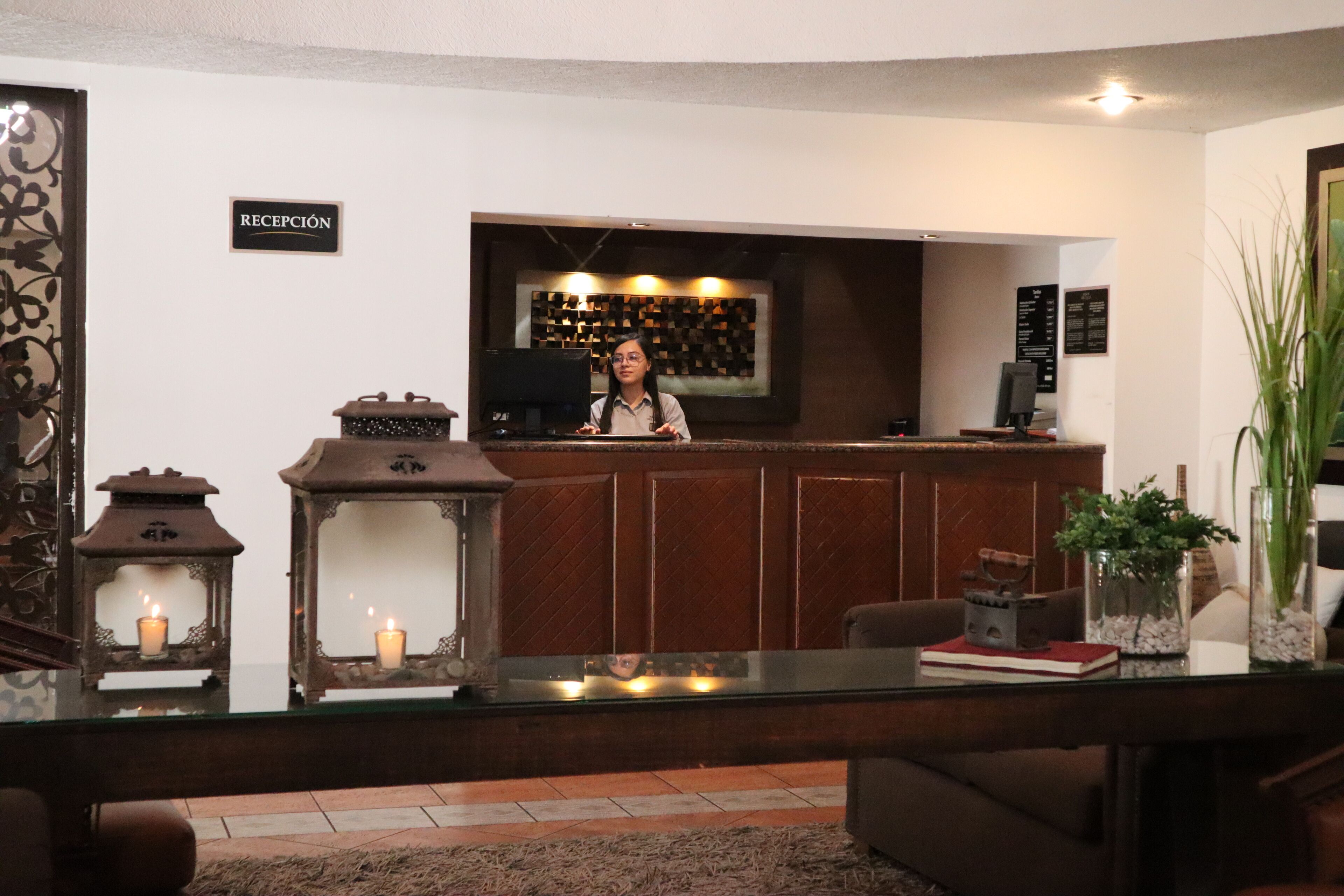 A reception area with a wooden counter, a sign reading "RECEPCIÓN", and a bar with a menu. There are two lit lanterns, a sofa, and a coffee machine on the counter.