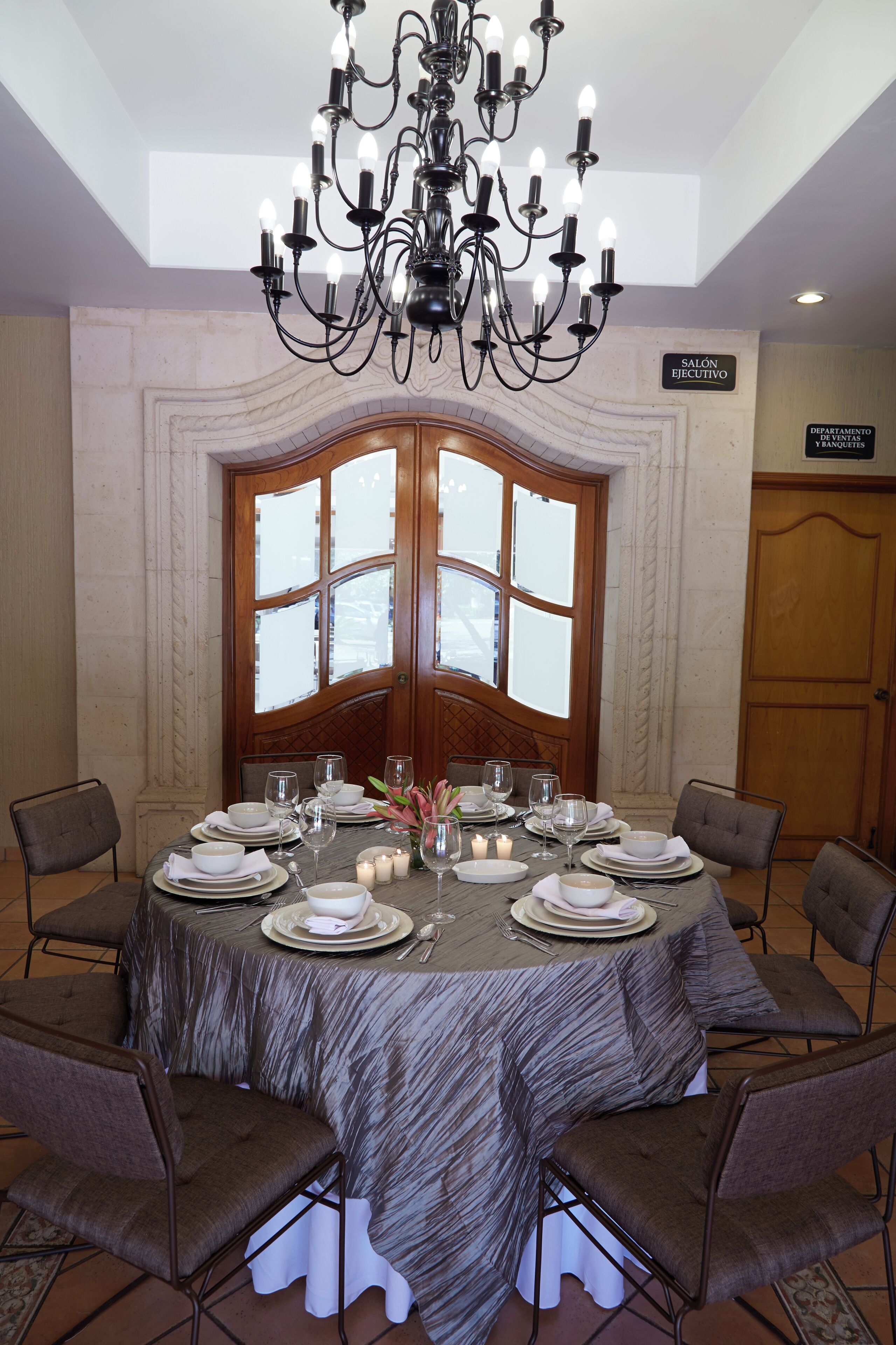 A dining room with a round table set for six, surrounded by six chairs. A chandelier hangs from the ceiling, and a large window with multiple panes is visible behind the table.