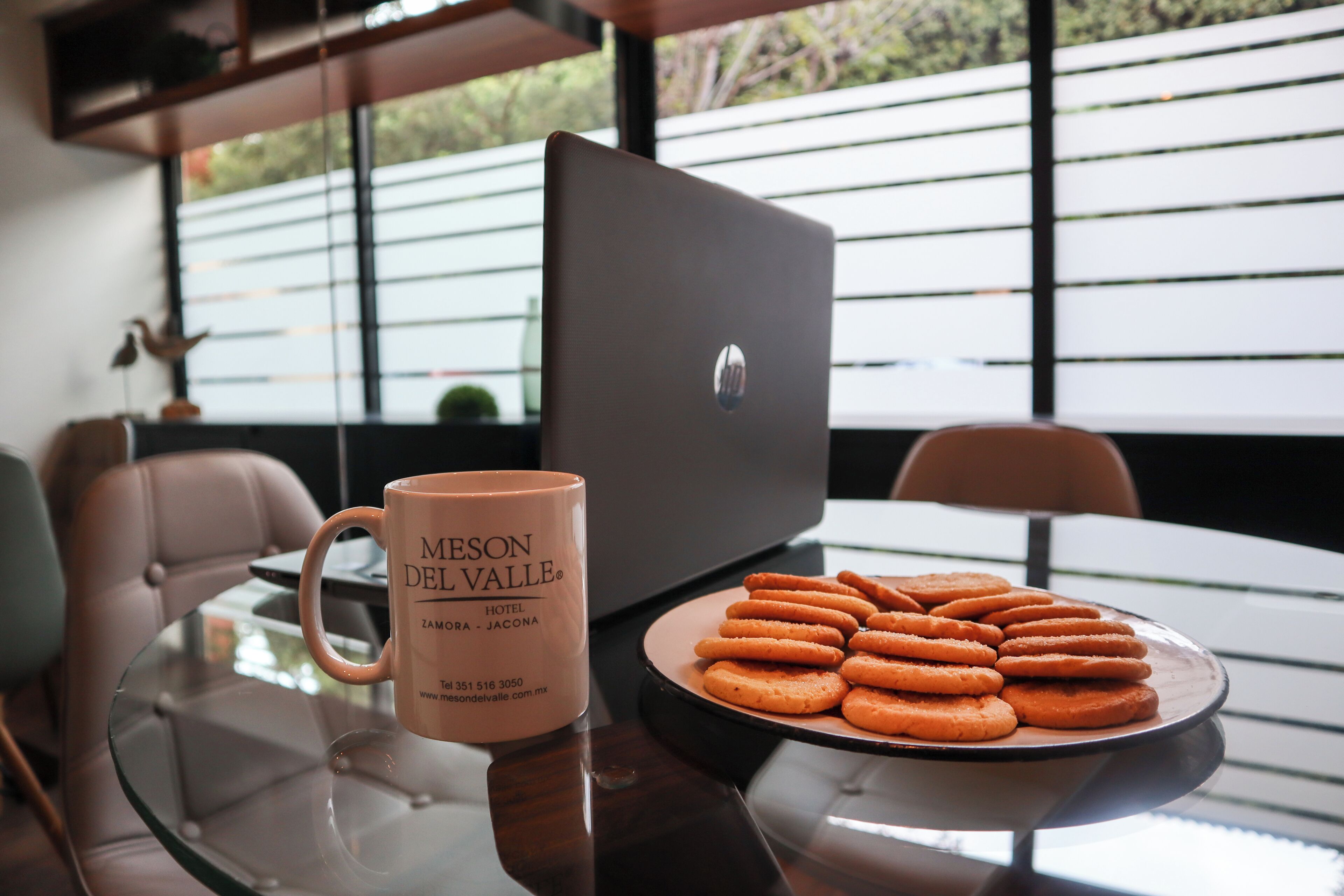 Une table en verre avec une tasse et une assiette de biscuits, un ordinateur portable et une chaise, dans une pièce moderne dotée de grandes fenêtres.