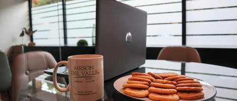 Une table en verre avec une tasse et une assiette de biscuits, un ordinateur portable et une chaise, dans une pièce moderne dotée de grandes fenêtres.