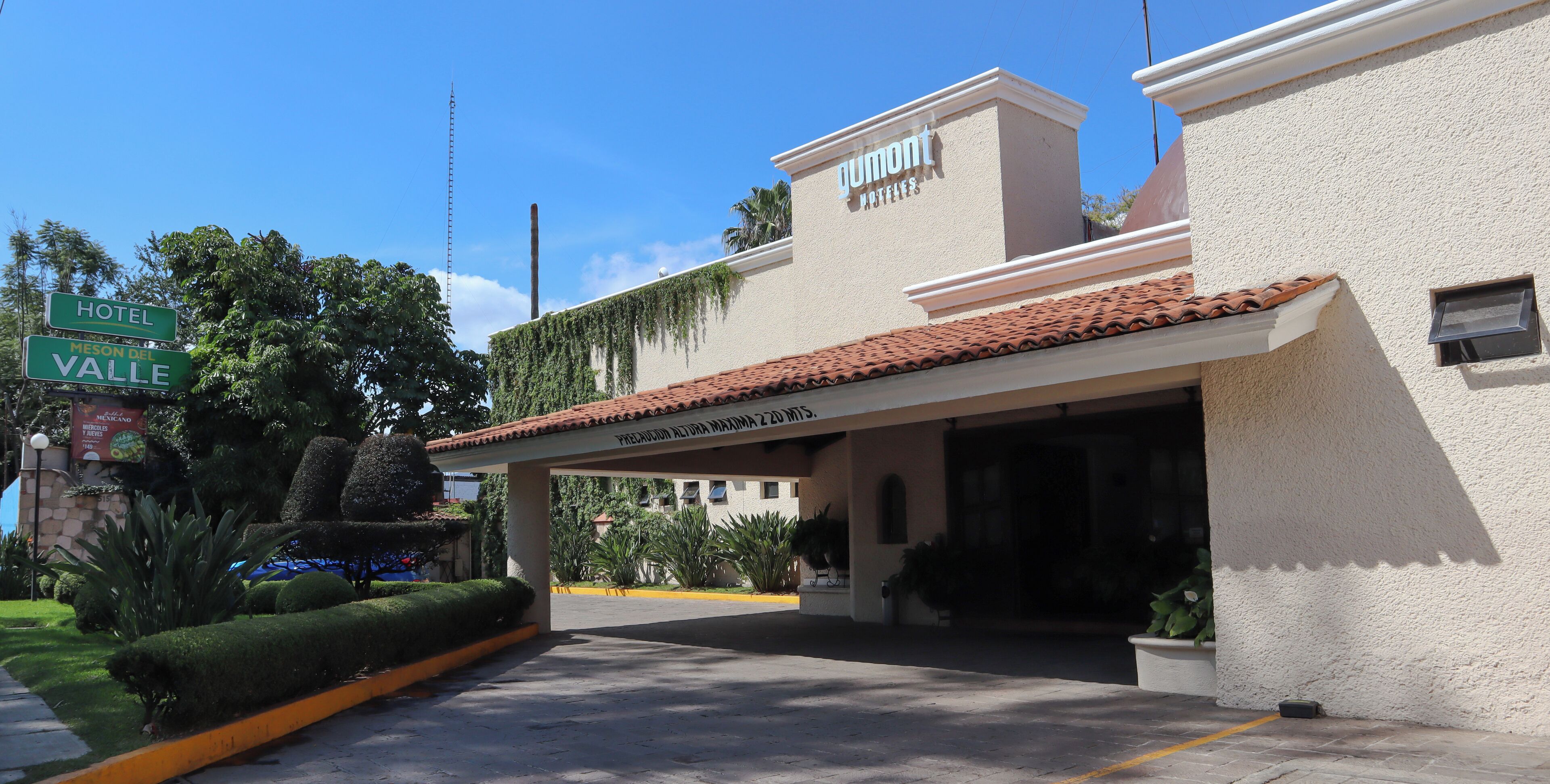 Entrada de hotel com galeria coberta, placa indicando 'Hotel Meson Del Valle', e um edifício com a sinalização 'Gumont Hotels'.