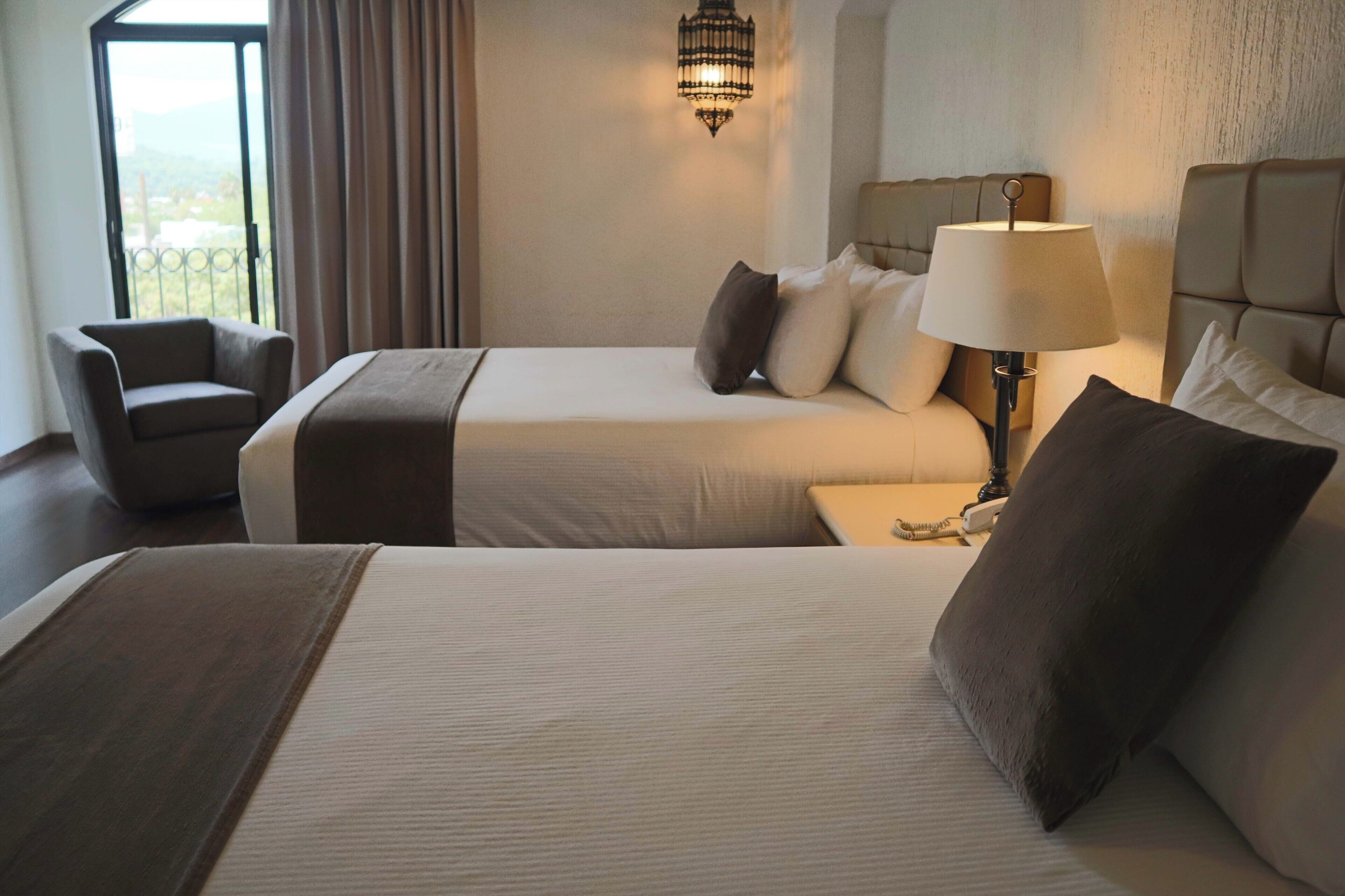 A hotel room with two beds, a chair, a lamp, and a view of the outdoors.