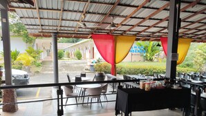 An outdoor dining area with tables and chairs, a covered roof, and a view of the surrounding greenery and buildings.