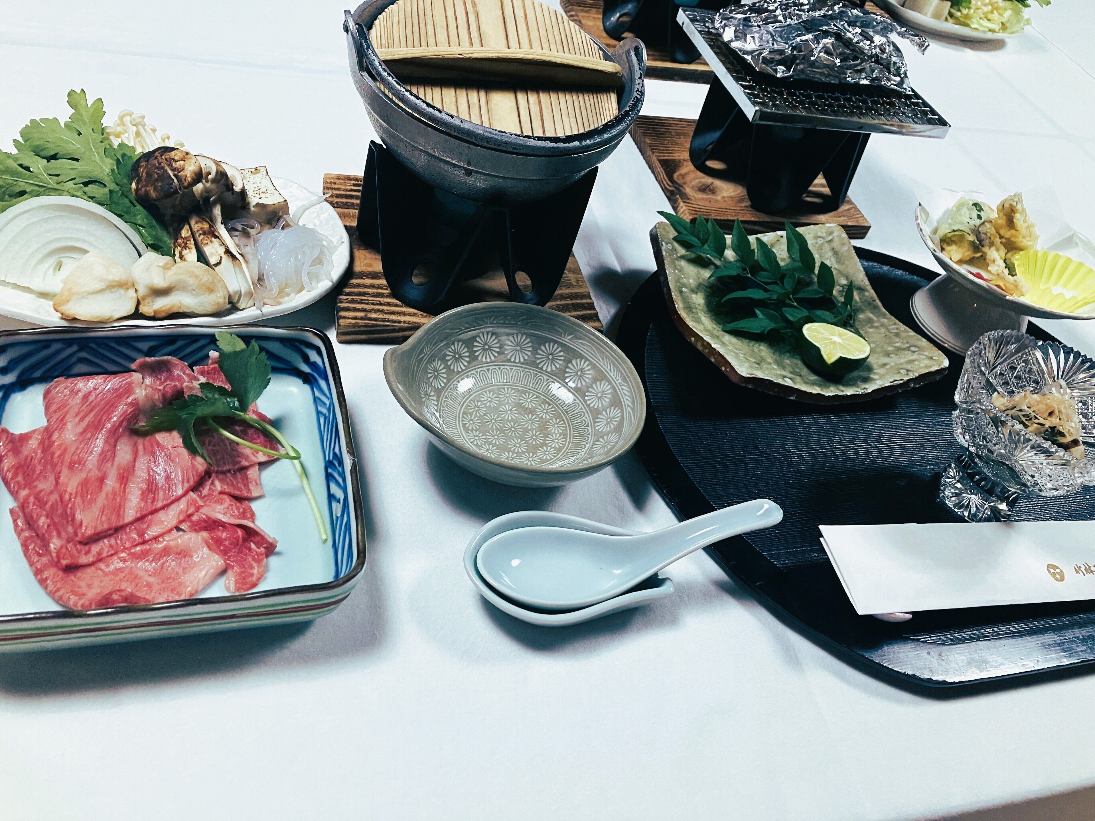 A table set with various dishes, including raw meat, vegetables, and a hot pot.