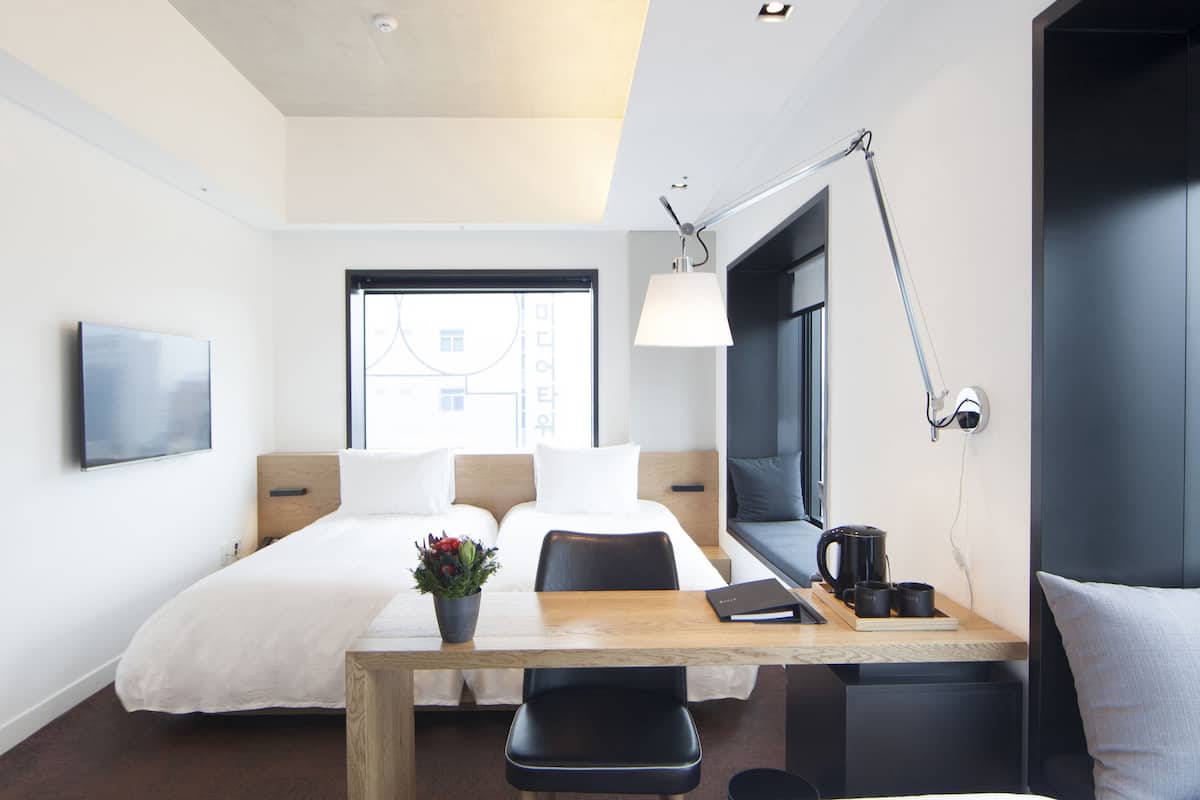 Ett hotellrum med två sängar, ett skrivbord med en stol, en TV och ett fönster med utsikt över byggnader.