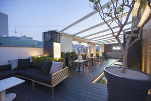 En takterrass med modern design, med en sittgrupp bestående av en soffa och ett bord, omgiven av planterade växter och ett stort träd.