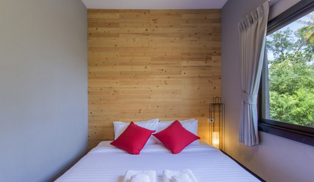 A bedroom with a wooden headboard, white bedding, and a view of greenery through a window.