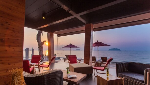 A covered outdoor seating area with red chairs and tables, overlooking a body of water and city skyline. - Lipda Resort (Prachuap Khiri Khan)