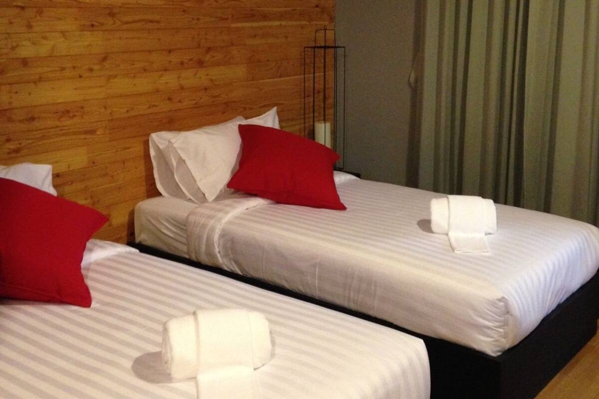 Two beds with white linens and red pillows, a wooden wall, and a green curtain.