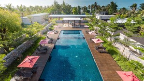 A rectangular swimming pool with a wooden deck, surrounded by lounge chairs and umbrellas, set against a backdrop of palm trees and a view of the sea. - Lipda Resort (Prachuap Khiri Khan)