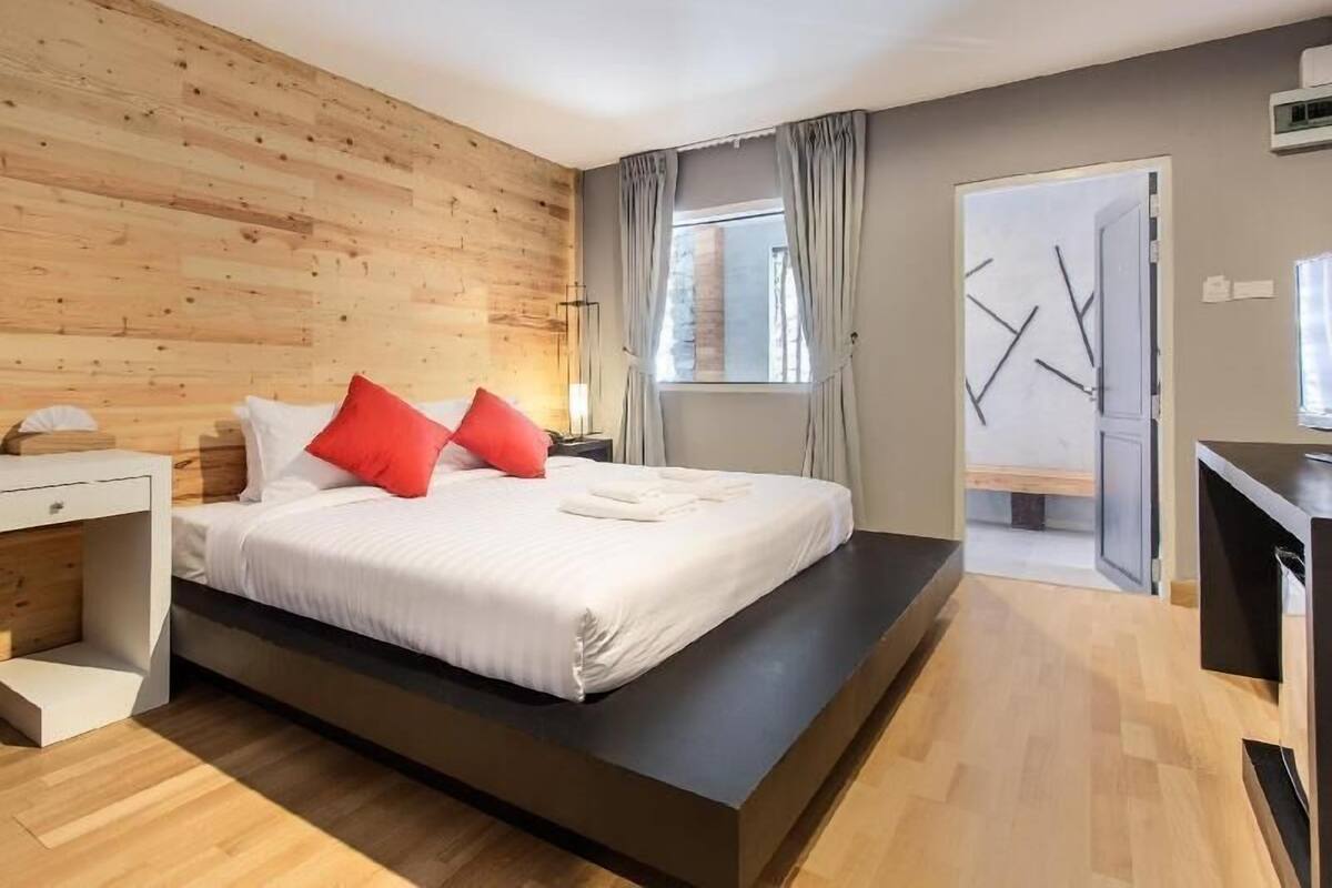 A modern bedroom with a wooden wall, a bed with white bedding and red pillows, a bedside table, a nightstand, a window with curtains, and a door with a mirror.