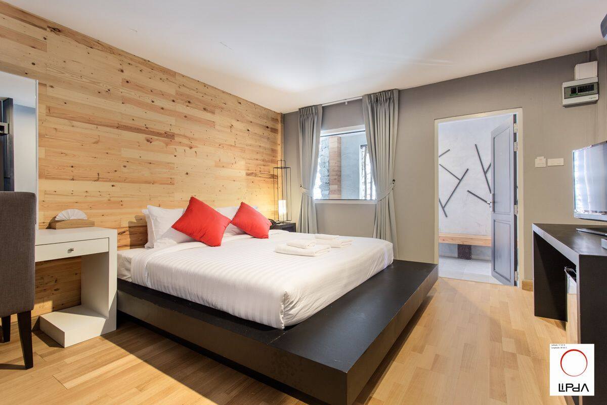 A modern bedroom with a wooden wall, a bed with white bedding and red pillows, a desk, and a TV.