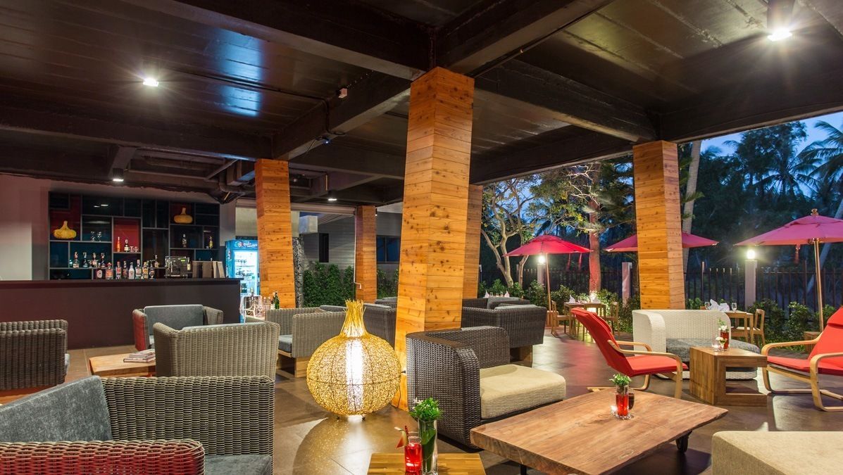 A modern outdoor seating area with wicker furniture, a bar in the background, and red umbrellas providing shade.