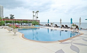 Ein Poolbereich mit Liegestühlen und Sonnenschirmen, Blick auf das Meer und ein Gebäude im Hintergrund.