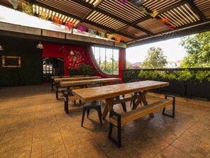 An outdoor dining area with wooden tables and benches, a red wall with floral artwork, and a view of greenery and buildings in the background.