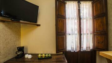 A room with a television mounted on the wall, a wooden desk with a coffee maker and decorative items, and a window with wooden shutters.