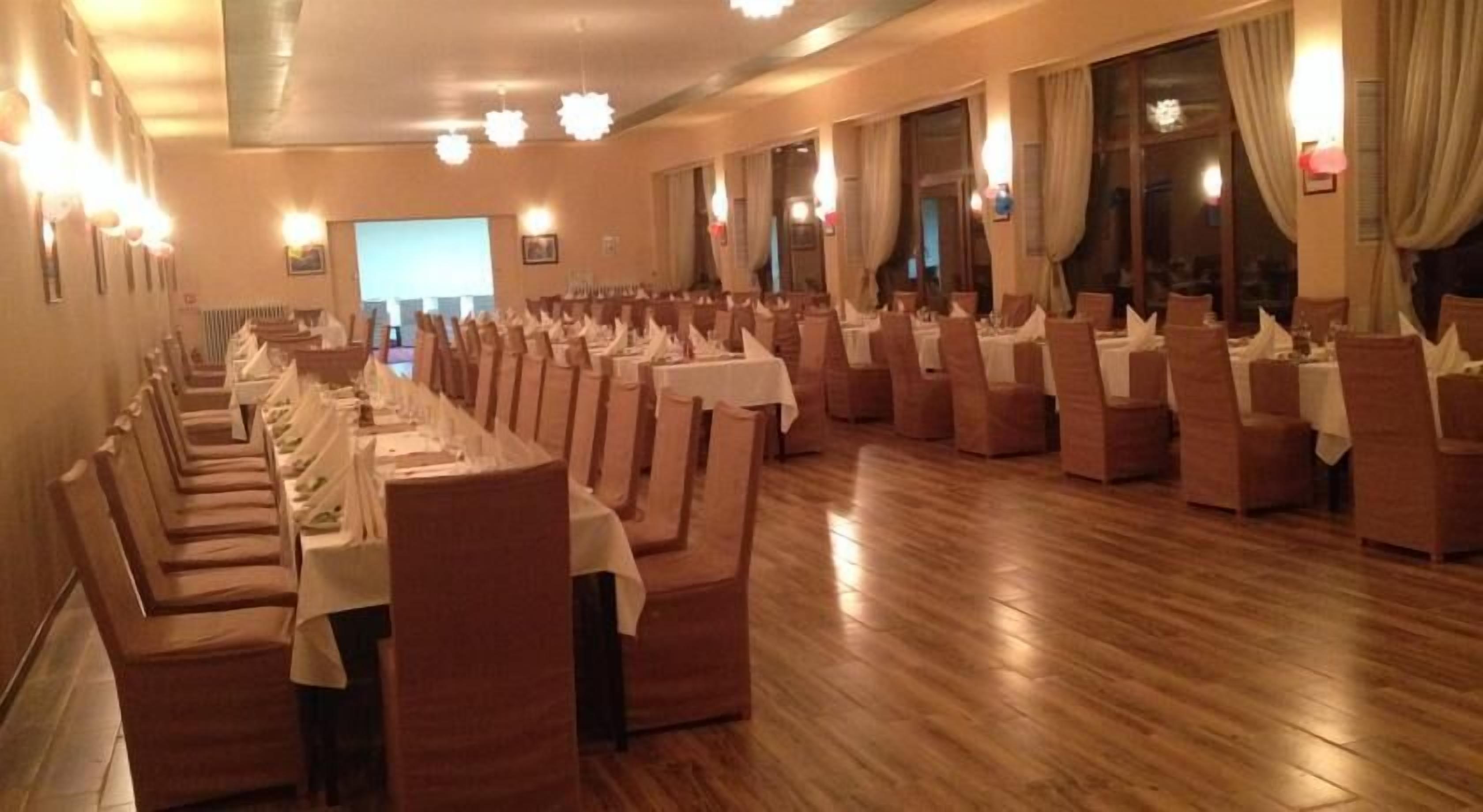 A banquet hall with rows of tables and chairs, wooden flooring, and large windows with curtains.
