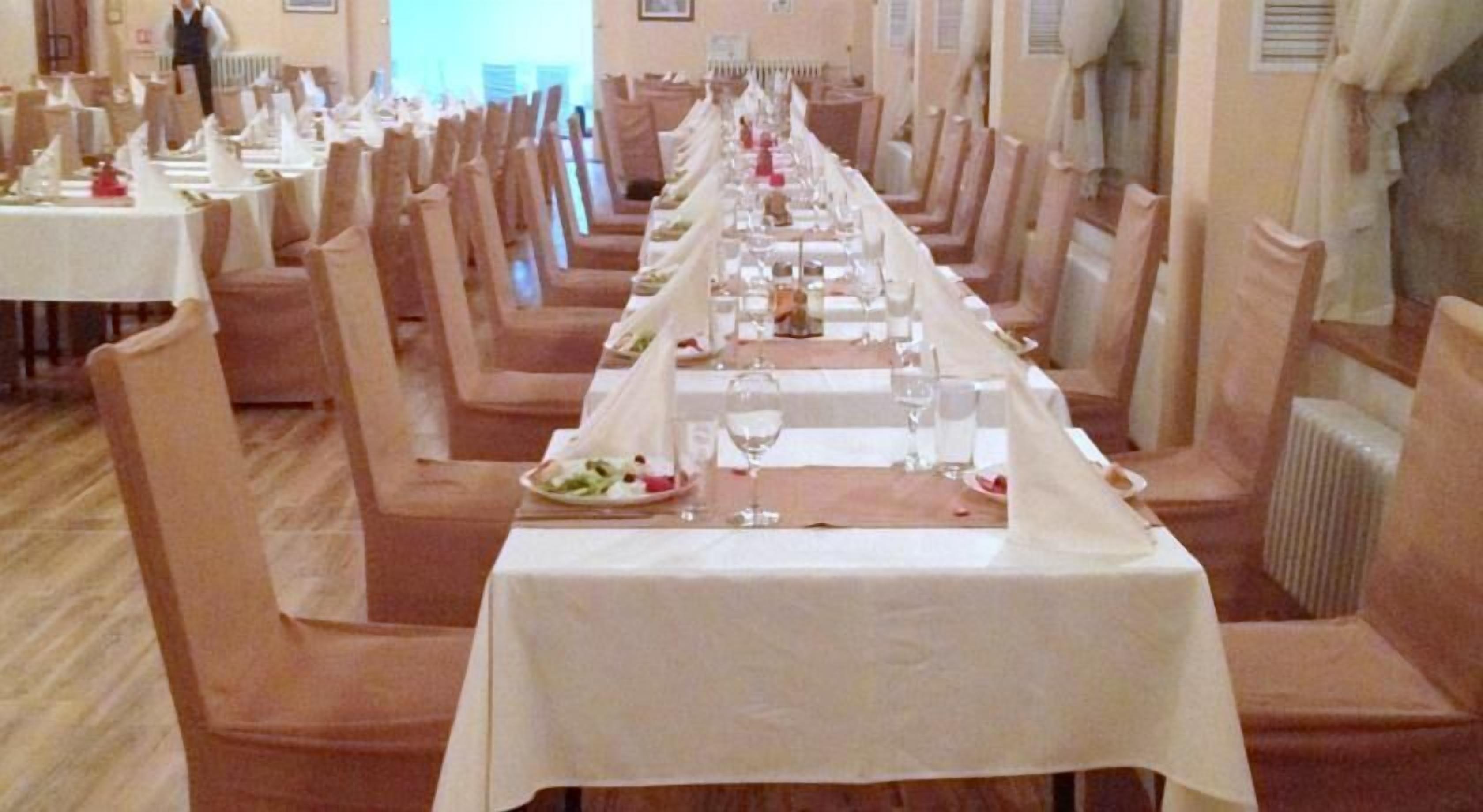 A long dining table with white tablecloths and chairs, set up in a room with wooden floors and beige walls.