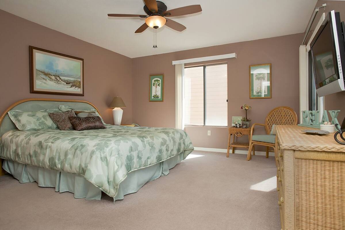 A bedroom with a bed, a ceiling fan, a TV, and a window.