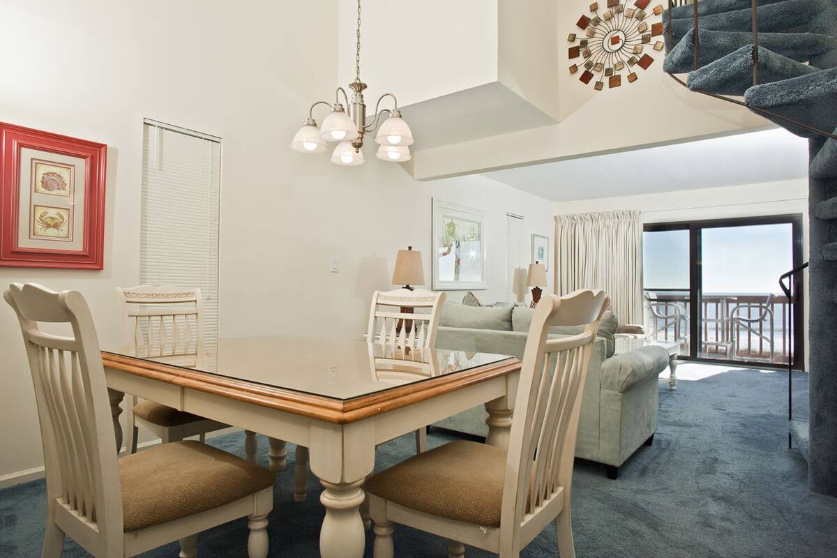 A dining area with a glass-top table, white chairs, and a chandelier. There is a balcony with a view of the ocean.