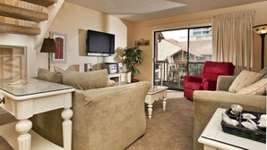 A living room with a sofa, armchair, and a red chair. There is a television mounted on the wall, a plant, and a window with curtains.