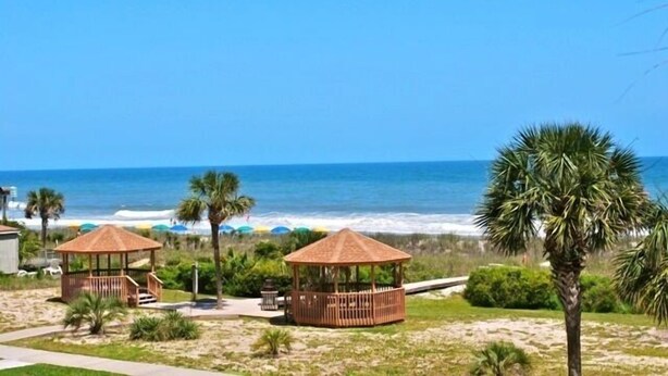 A beach view with two wooden gazebos, a palm tree, and the ocean in the background.