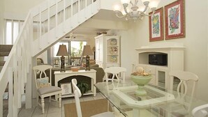 A spacious living room with a glass dining table, white furniture, a fireplace, and a staircase.