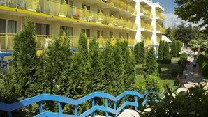 Un hotel de varios pisos con balcones, una torre blanca central y un jardín ajardinado con cerca azul.