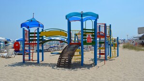 Un parque infantil en la playa con tobogán, estructuras para escalar y una zona sombreada.