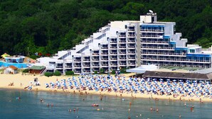 Un hotel frente a la playa con varios balcones, una playa de arena y numerosas sombrillas.