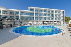 Un hotel moderno con una piscina grande, mobiliario exterior y un edificio de varios pisos con balcones.