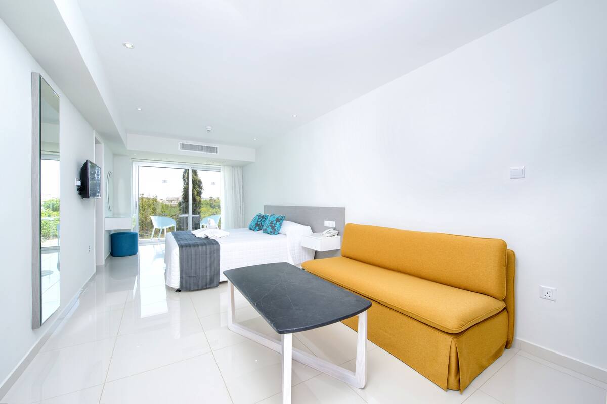 A modern, bright room with a bed, a sofa, a small table, and a large window offering a view of greenery outside.
