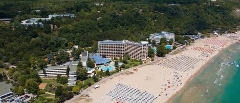 An aerial view of a coastal resort with multiple hotel buildings, a large sandy beach with numerous umbrellas, and a clear view of the ocean.