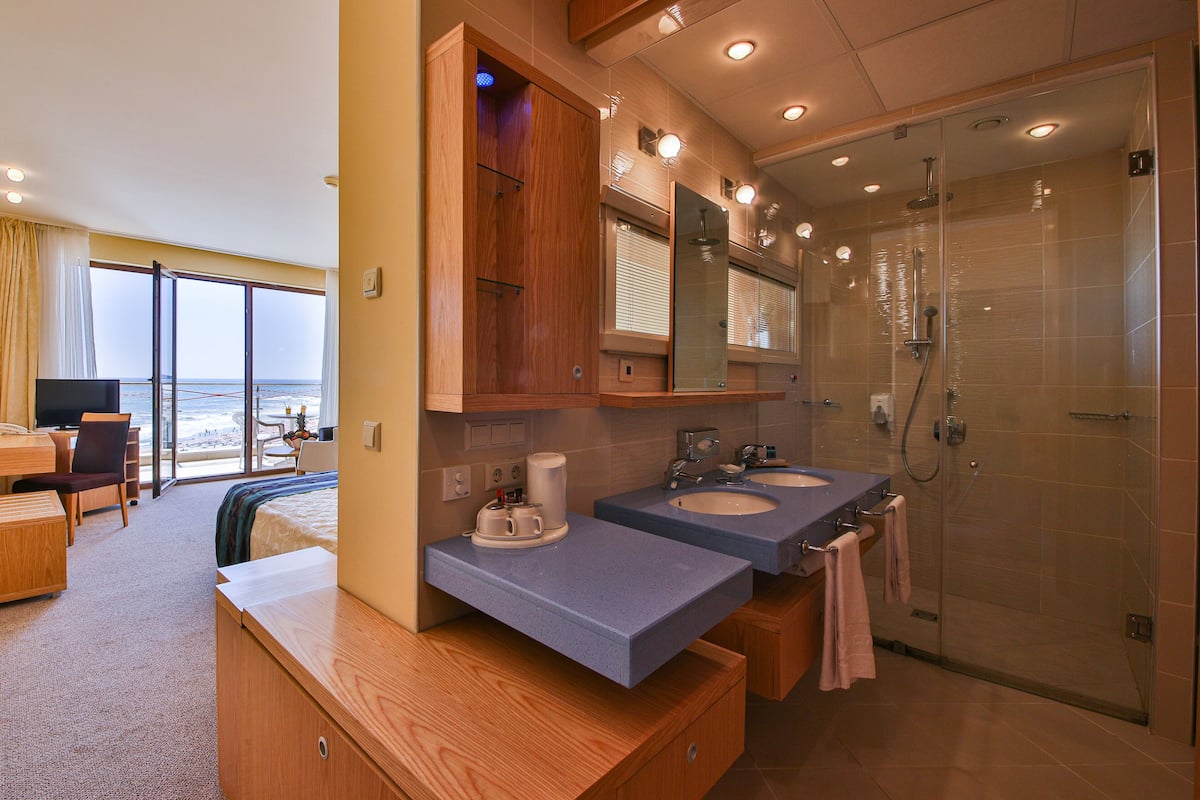 A hotel room with a balcony, a desk with a chair, a bed, a bathroom with two sinks and a shower.