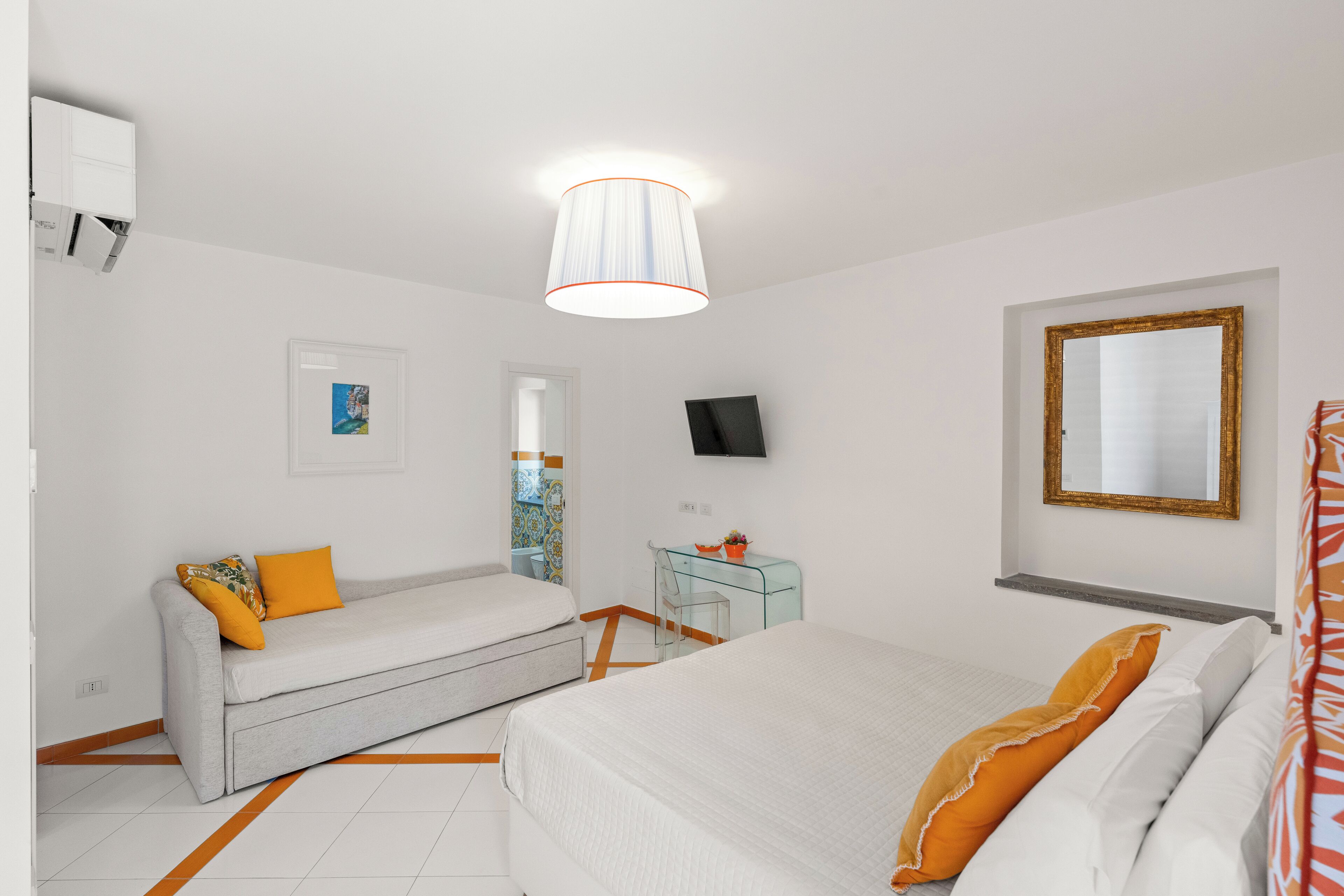 Quarto de hotel com cama, sofá, escrivaninha com cadeira, televisão e espelho.