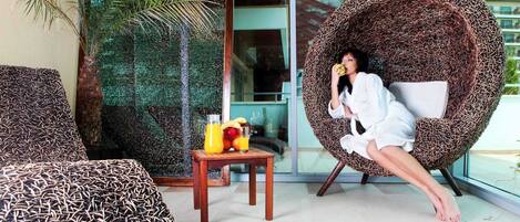 A modern interior with a large, round, woven chair, a patterned sofa, and a small table with fruit and a glass.
