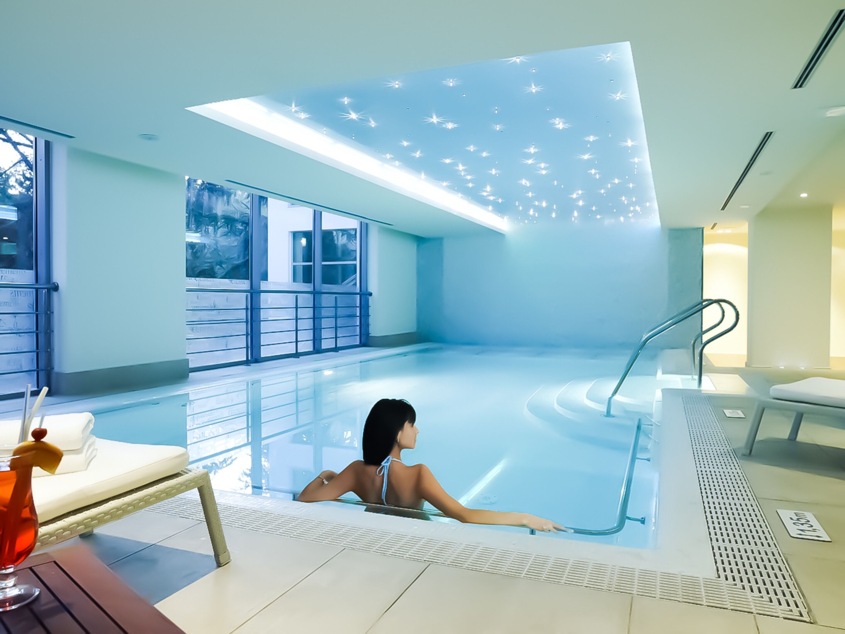 A modern indoor swimming pool with a glass wall, a person in the water, and a lounge area with a drink.